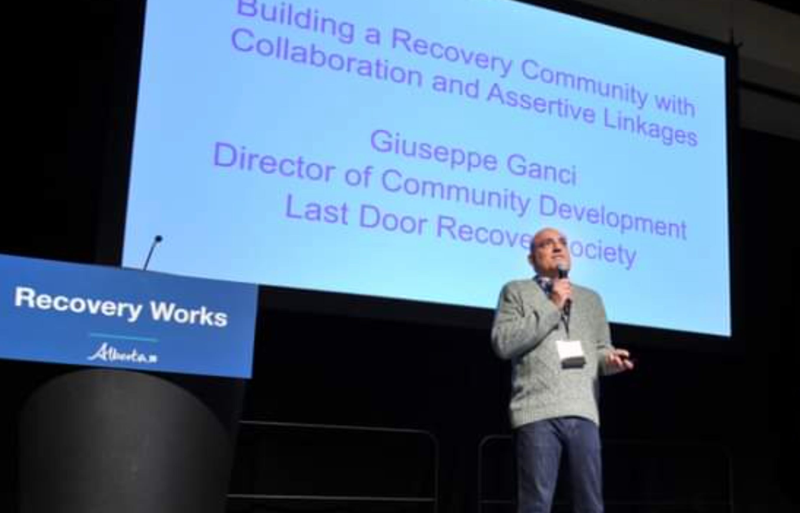 Guiseppe Ganci stands on a stage with a presentation title slide behind him, which reads building a recovery community with collaboration and assertive linkages, beside a podium that reads recovery works