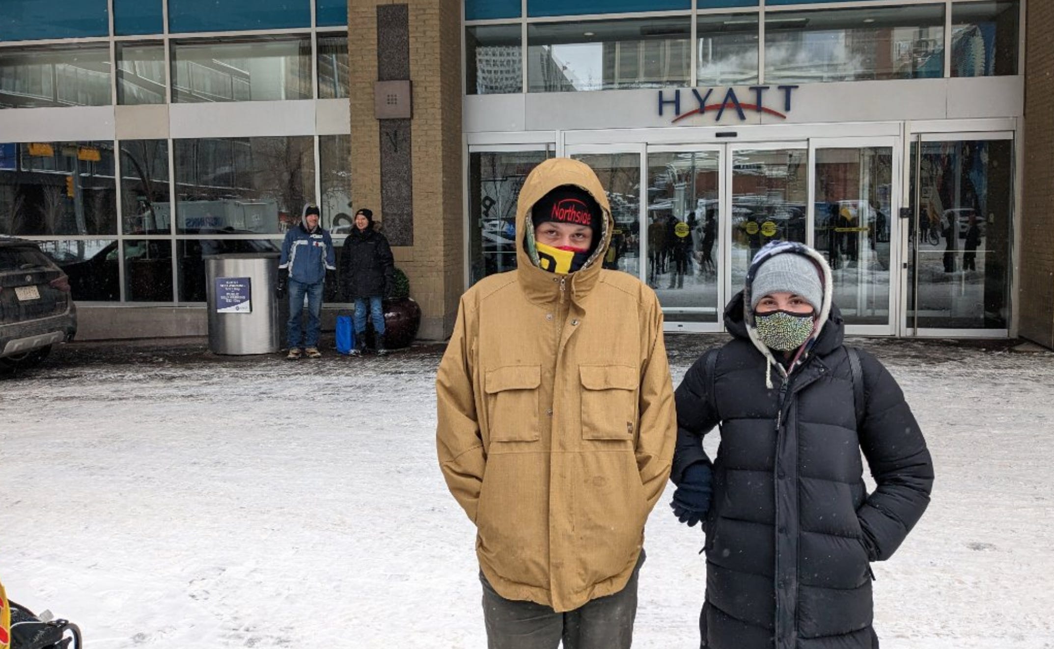 Two people stand in the foreground bundled up head to toe including face coverings, with two characters in the background beside the entrance to the Hyatt Regency in downtown Calgary. Snow is on the ground all around