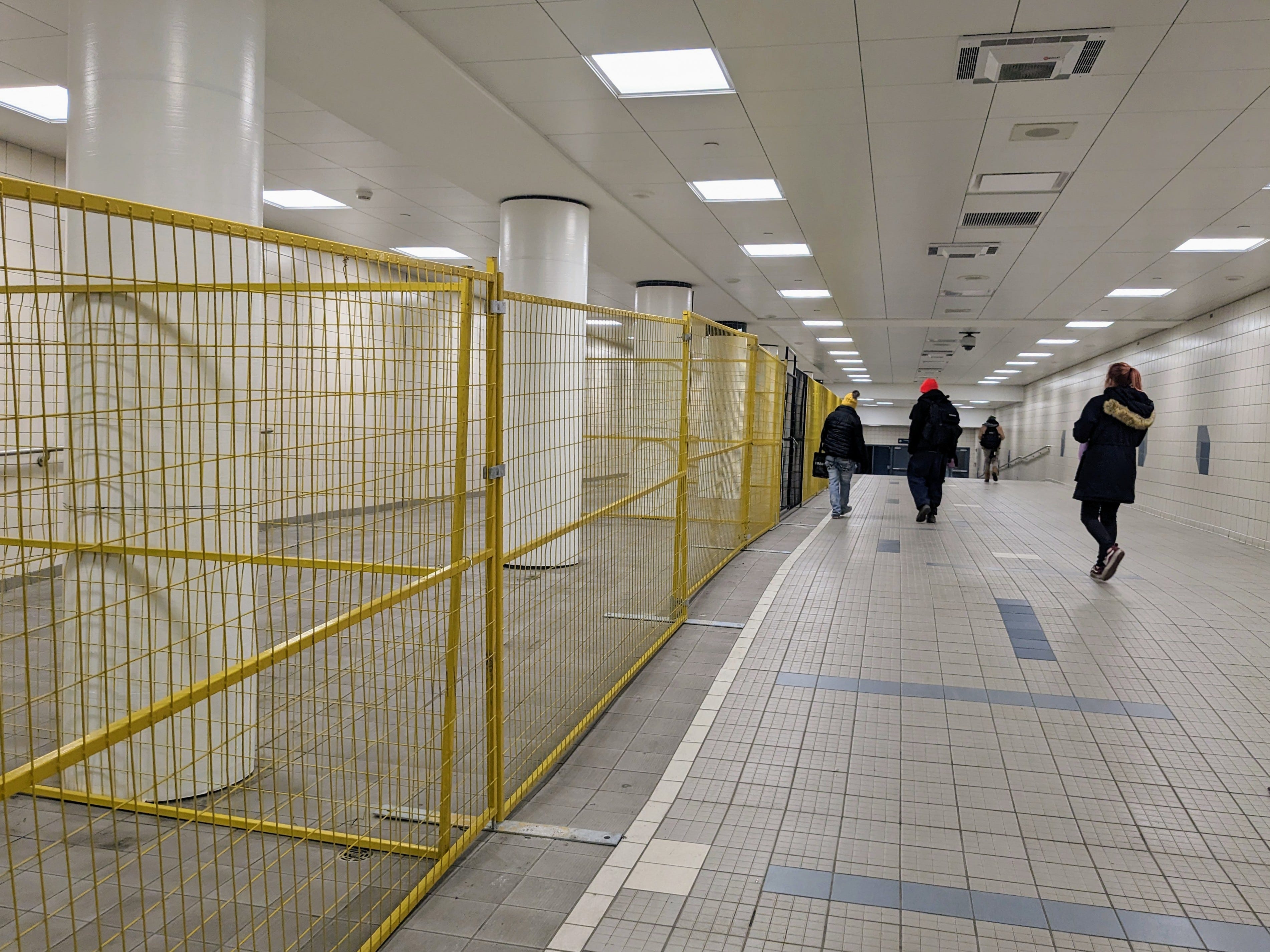 A transit tunnel in Edmonton is underground, showing people walking through alongside a yellow steel fence that closes off an open area in the tunnel, where on house people used to congregate and keep warm. Fluorescent lights beam overhead. The photo has a sense of coldness and Cavernous space
