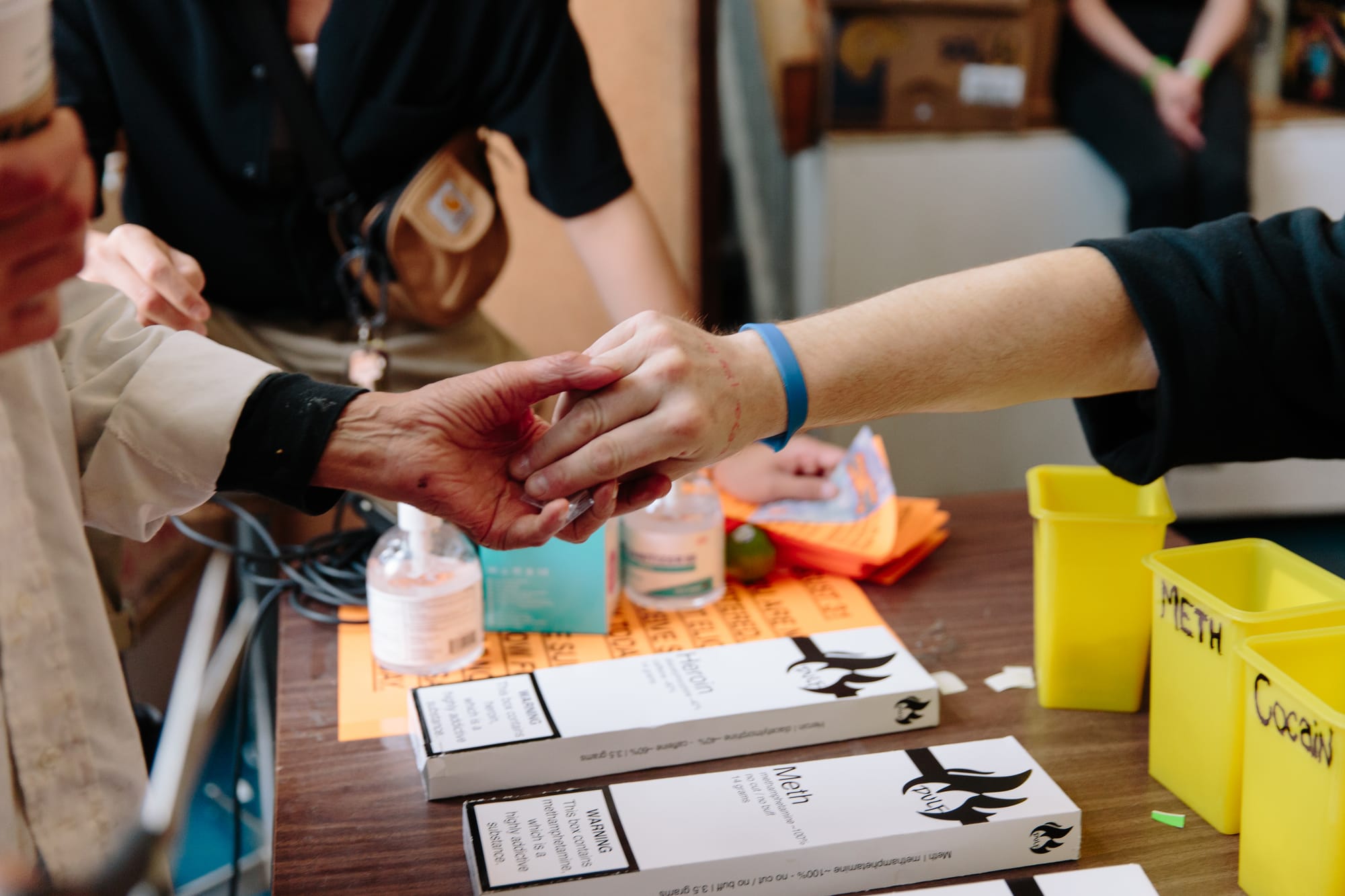 Two hands meet over a table, giving and receiving a regulated drug in a small baggie. Boxes indicating regulated heroin and meth are visible nearby.