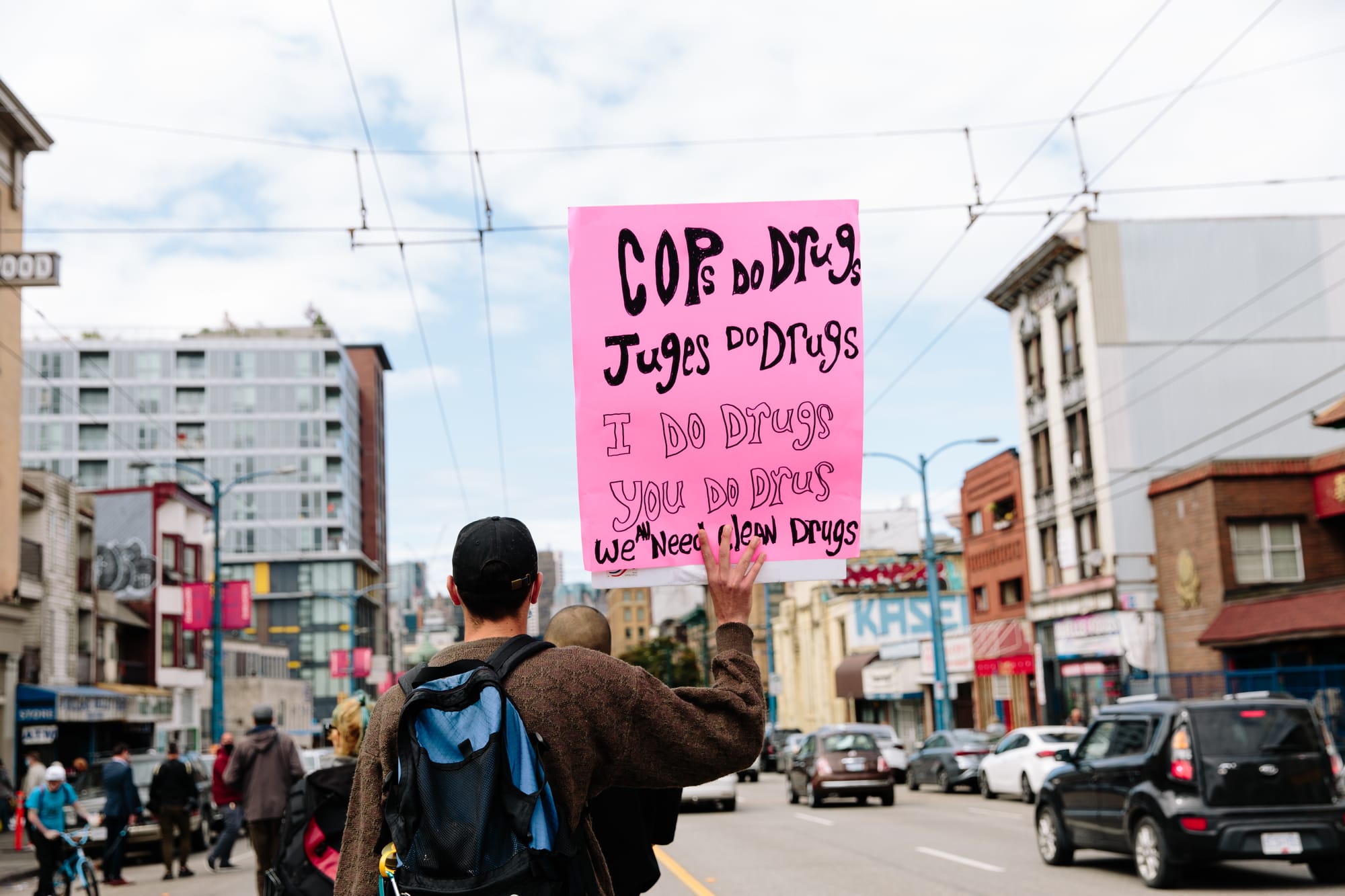 Rear view of a person in a brown sweater and blue backpack holding up a pink sign that reads "Cops do drugs. Judges do drugs. I do drugs. You do drugs. We need new drugs." Down what appears to be Hastings Street in Vancouver.