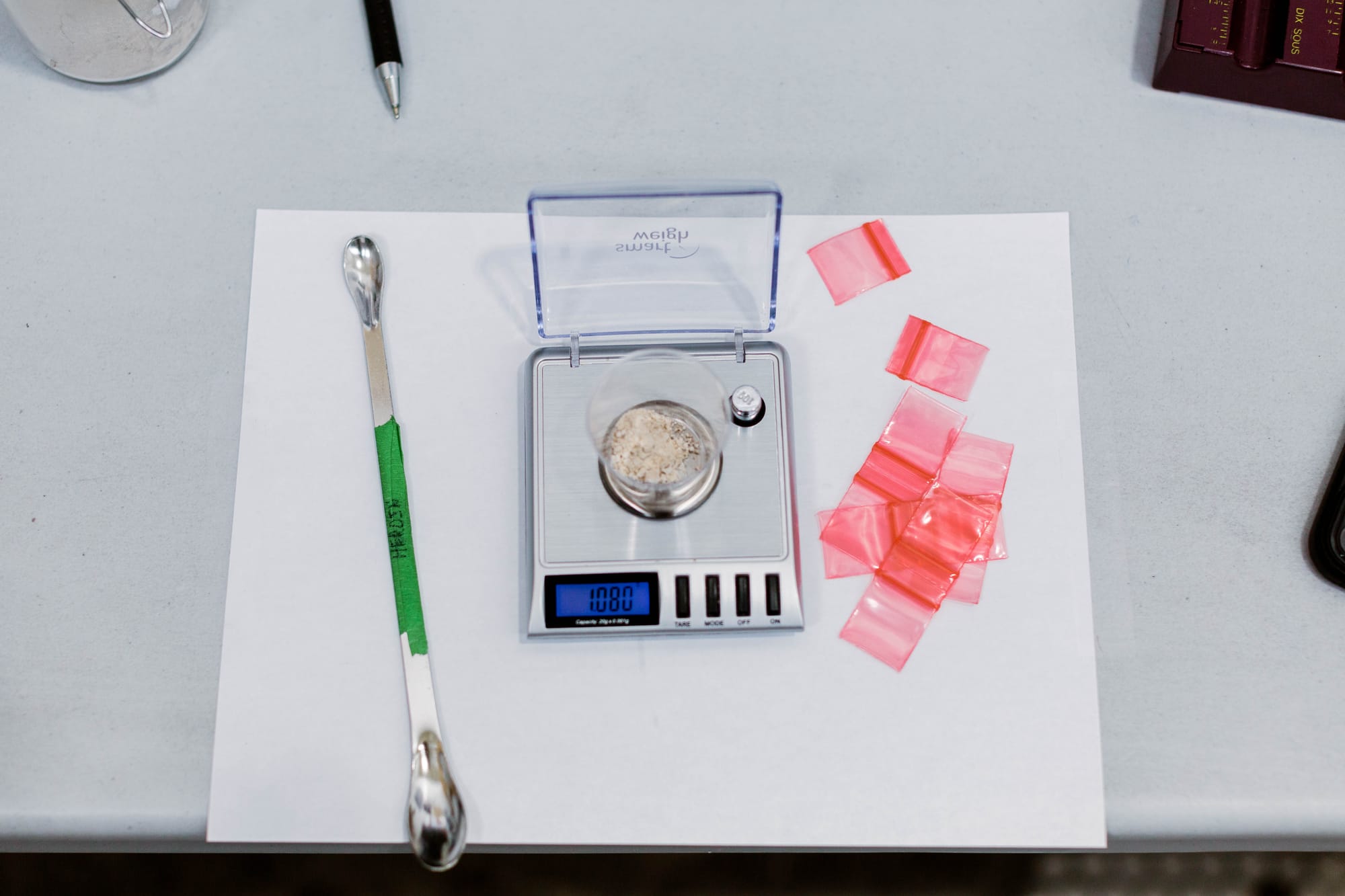 Aseptic scene of a scoopula (lab spoon) and weigh scale with red plastic ziploc bags beside it. A powder substance is in a cup on the scale.