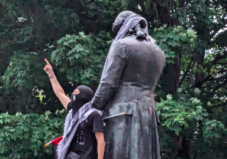 A person wearing a keffiyeh points a finger at the sky while standing next to a statue of a man, on which a keffiyeh was hung around the neck.