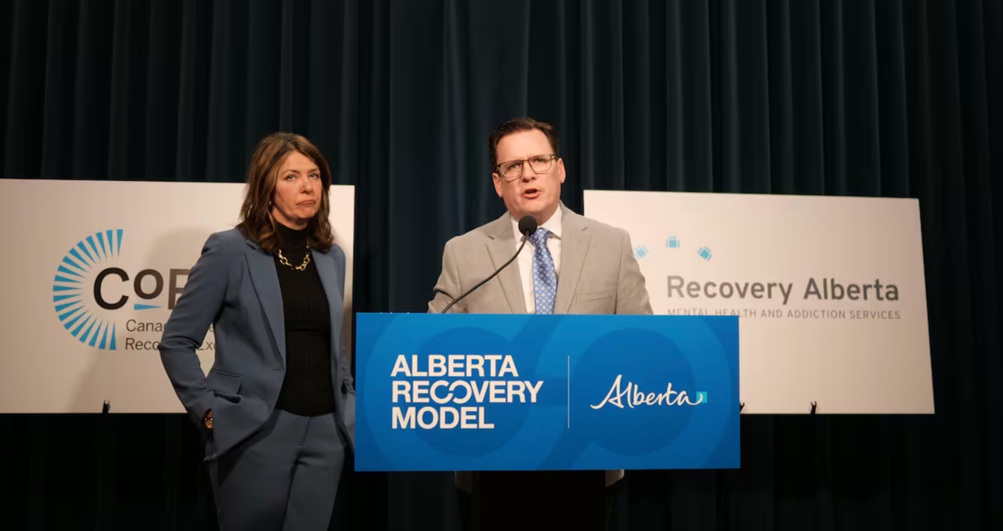 A man and a woman in formal attire stand speaking at a podium that says "Alberta Recovery Model." The man's mouth is open as if speaking and the woman looks somewhat glum. The curtains behind them are black and two white signs say "CoRE" and "Recovery Alberta."