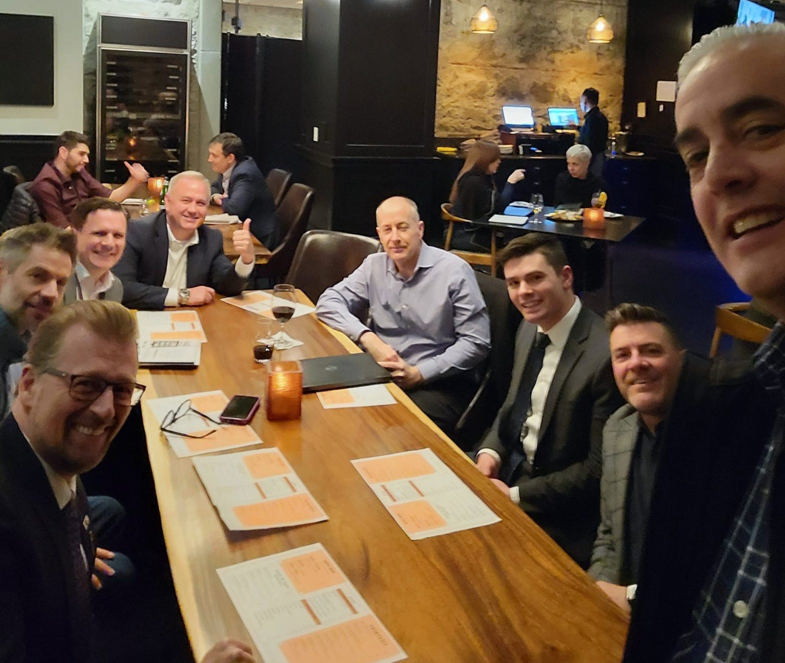 A group of white men ranging in age from twenties to sixties sit grouped around a long wooden table, with conference programs in front of them like placemats. One of them gives a thumbs-up. 