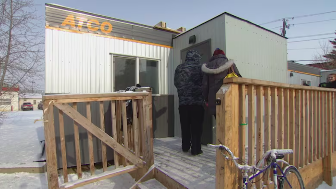 Two people stand outside a closed door of a portable Atco trailer in the winter.