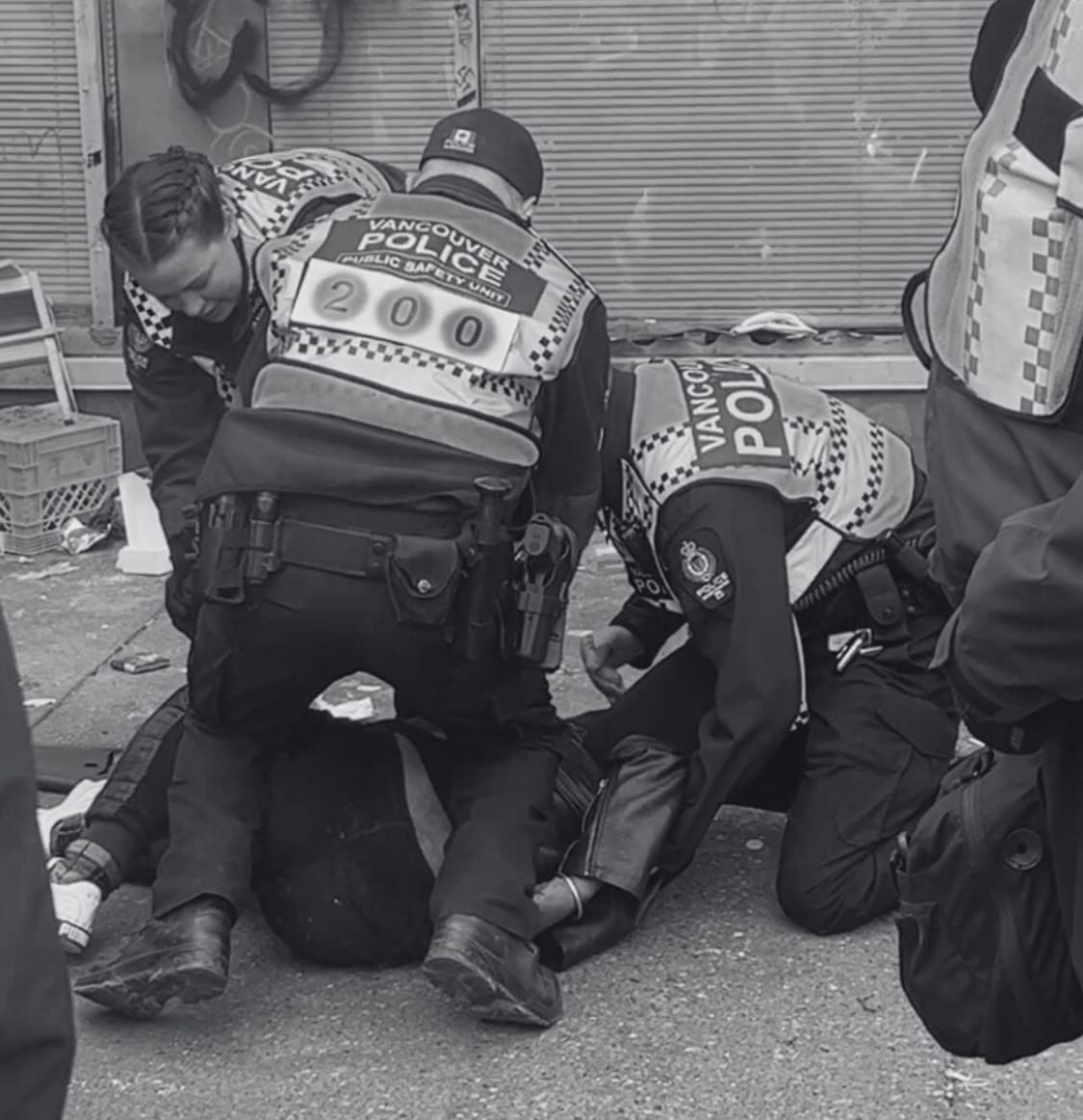 3 police officers kneel on and surround a person lying prone on the sidewalk with one arm at a painful-looking angle.