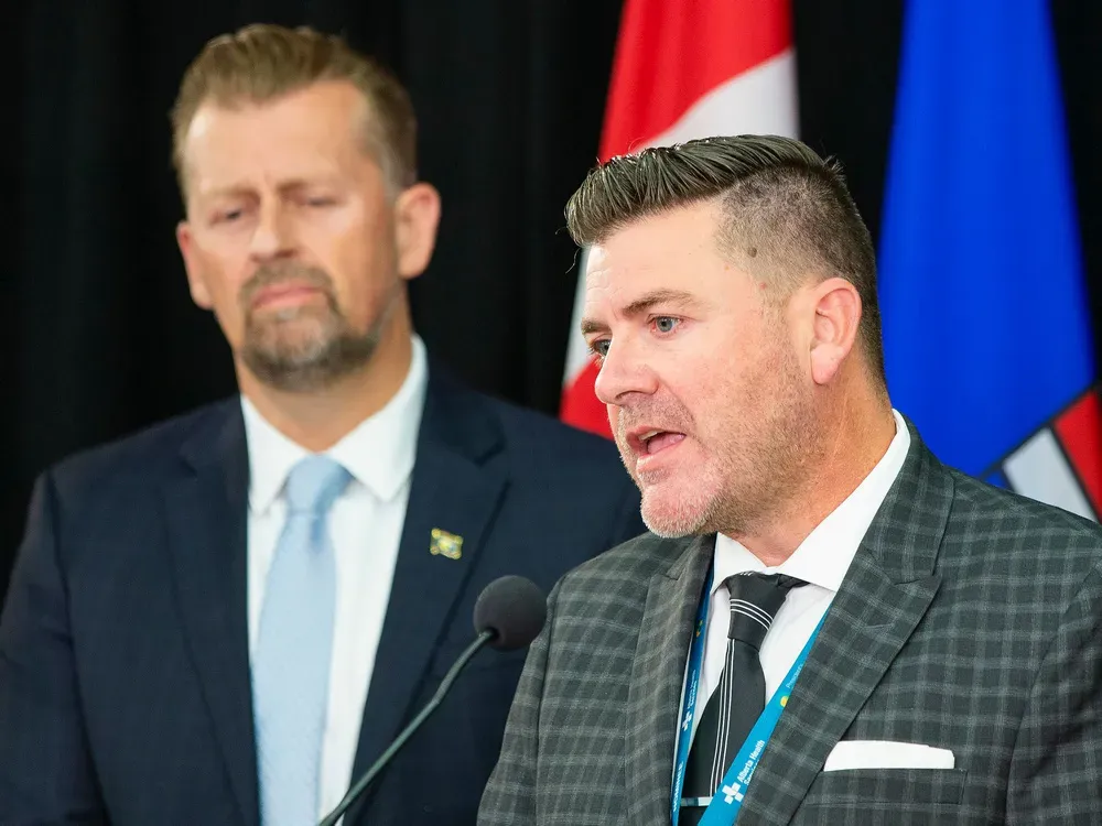 A man in a two-tone tartan suit jacket and heavily gelled hair speaks into a microphone. Another man lurks in the background.