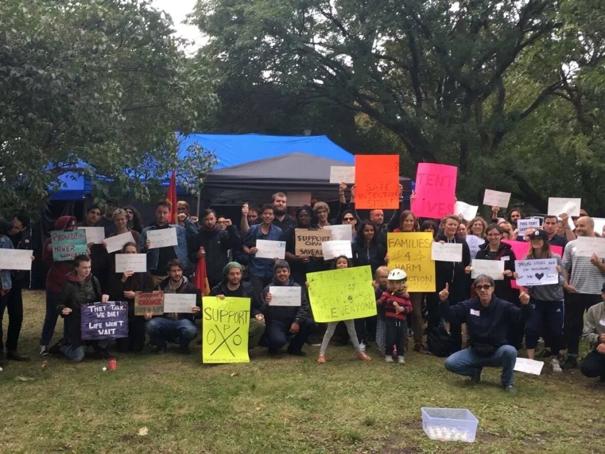 A group of around fifty people pose with signs declaring their support for overdose prevention sites, in a forested field.