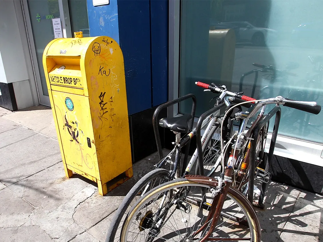 A yellow needle drop box sits in front of the windows and security door of a building, beside some parked bicycles. 