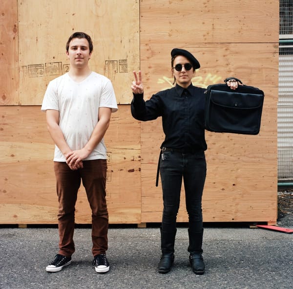 Two people stand face the camera in front of a plywood wall. One holds up a peace sign.