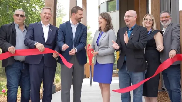 Seven people smile at the camera and at one another while clapping in a row in front of a red ribbon being cut.