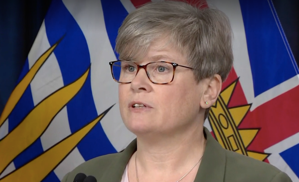 A woman with glasses in front of a BC flag speaks to a crowd at a press conference.