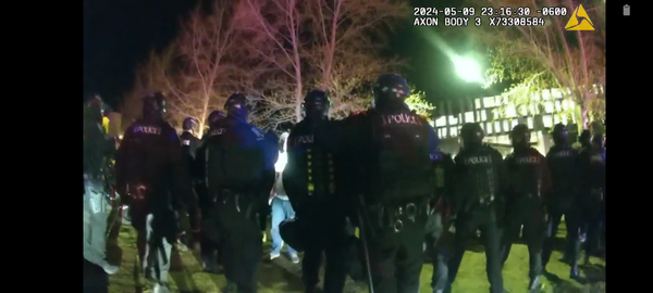 Fish-eye view from a body-worn police camera, showing 15 officers in riot gear facing a protest, from behind.