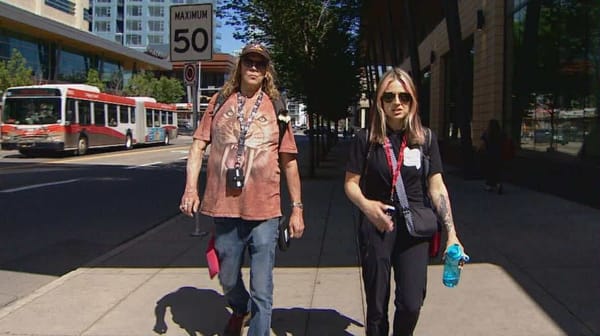 A tall man with long blonde hair and a woman with blonde and pink hair walk on a city street wearing backpacks.