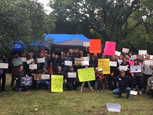 A group of around fifty people pose with signs declaring their support for overdose prevention sites, in a forested field.